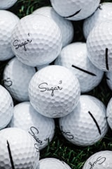 A collection of white golf balls with the word 'Sugar' printed on them in black script. The golf balls are stacked closely together on a surface with patches of green grass visible in the background.