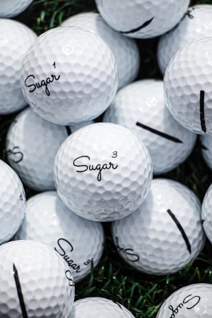 A collection of white golf balls with the word 'Sugar' printed on them in black script. The golf balls are stacked closely together on a surface with patches of green grass visible in the background.