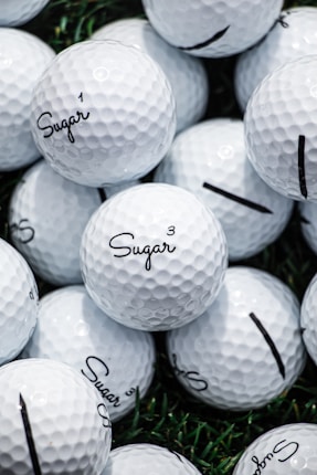 A collection of white golf balls with the word 'Sugar' printed on them in black script. The golf balls are stacked closely together on a surface with patches of green grass visible in the background.