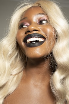 A person with bright, blonde hair and a wide smile. The makeup includes black lipstick and pinkish eyeshadow. The skin appears smooth and reflects the lighting, adding a glossy effect.