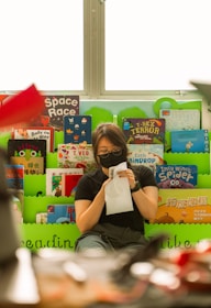 A person with glasses and a black mask is sitting in front of a colorful display of children's books, holding a piece of paper. The books have vibrant covers with titles such as 'Space Race,' 'I-Rex Terror,' and 'Incy Wincy Spider.' The background is bright and well-lit from a window above.
