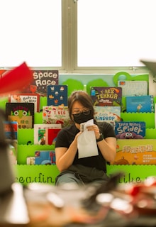 A person with glasses and a black mask is sitting in front of a colorful display of children's books, holding a piece of paper. The books have vibrant covers with titles such as 'Space Race,' 'I-Rex Terror,' and 'Incy Wincy Spider.' The background is bright and well-lit from a window above.