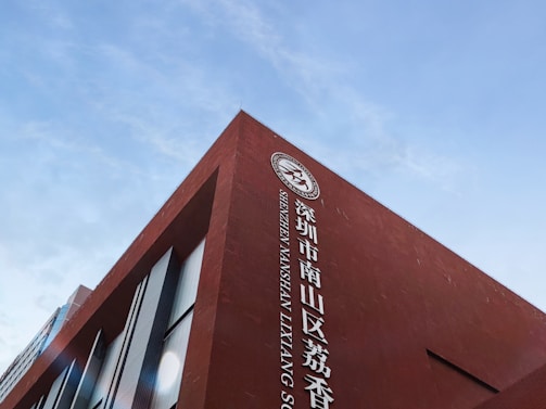 A red brick building with large white lettering in both Chinese and English, featuring the words 'Shenzhen Nanshan Laixiang'. A logo appears on the top portion of the wall. The sky is clear and blue, providing a contrast to the building's vibrant color.