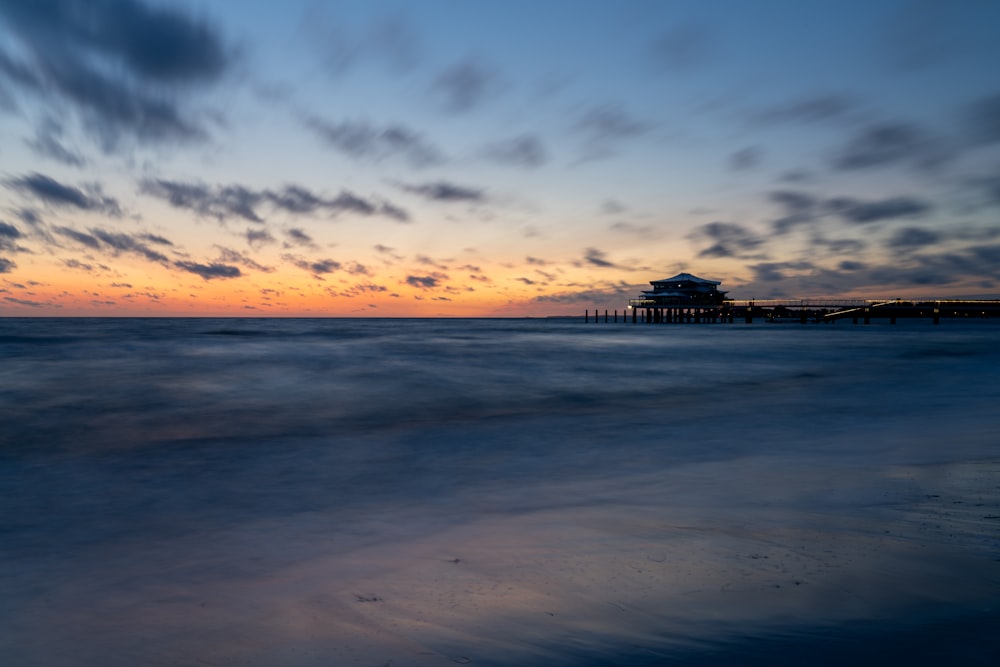 the sun is setting over the ocean with a pier in the distance