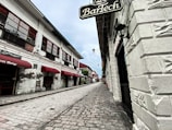 A cobblestone street lined with historical buildings featuring white brick walls and red awnings. The perspective leads down a narrow alleyway with traditional-style lanterns mounted on the walls. Signs with vintage-style fonts are visible, pointing to a heritage bakery and other businesses.