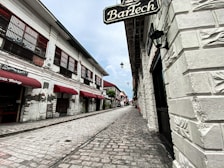 A cobblestone street lined with historical buildings featuring white brick walls and red awnings. The perspective leads down a narrow alleyway with traditional-style lanterns mounted on the walls. Signs with vintage-style fonts are visible, pointing to a heritage bakery and other businesses.