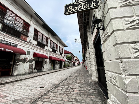 A cobblestone street lined with historical buildings featuring white brick walls and red awnings. The perspective leads down a narrow alleyway with traditional-style lanterns mounted on the walls. Signs with vintage-style fonts are visible, pointing to a heritage bakery and other businesses.