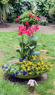 A vibrant garden scene with a large planter in the center filled with red, orange, and blue flowers. The background contains a lush green garden with more blooming flowers. A small decorative metal sculpture resembling a dog is placed in front of the planter.