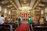 A richly decorated interior of a church with ornate columns, large statues, and colorful stained glass windows. The congregation, dressed in vibrant traditional attire, is seated on wooden pews, focusing on the altar. Floral arrangements and red drapery accentuate the space, highlighting the central religious figures.
