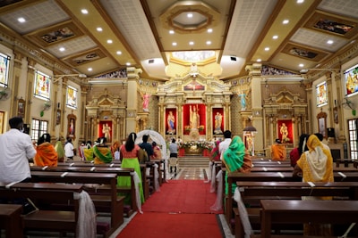 A richly decorated interior of a church with ornate columns, large statues, and colorful stained glass windows. The congregation, dressed in vibrant traditional attire, is seated on wooden pews, focusing on the altar. Floral arrangements and red drapery accentuate the space, highlighting the central religious figures.