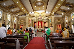 A richly decorated interior of a church with ornate columns, large statues, and colorful stained glass windows. The congregation, dressed in vibrant traditional attire, is seated on wooden pews, focusing on the altar. Floral arrangements and red drapery accentuate the space, highlighting the central religious figures.