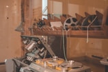 A vintage reel-to-reel tape machine is positioned in a room with a wooden shelf above it, holding several film reels. The machine appears to have multiple dials and metallic components. A retro lamp is attached to the wall beside the machine, casting soft light onto the scene.