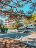 A modern residential building surrounded by lush greenery and trees. There are palm trees and neatly trimmed hedges in front of the building. Cars are parked along the street in front of the building, with a bicycle rack in the foreground. The sky is clear and blue, indicating a bright day.