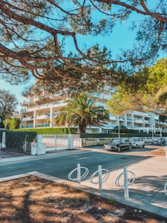A modern residential building surrounded by lush greenery and trees. There are palm trees and neatly trimmed hedges in front of the building. Cars are parked along the street in front of the building, with a bicycle rack in the foreground. The sky is clear and blue, indicating a bright day.