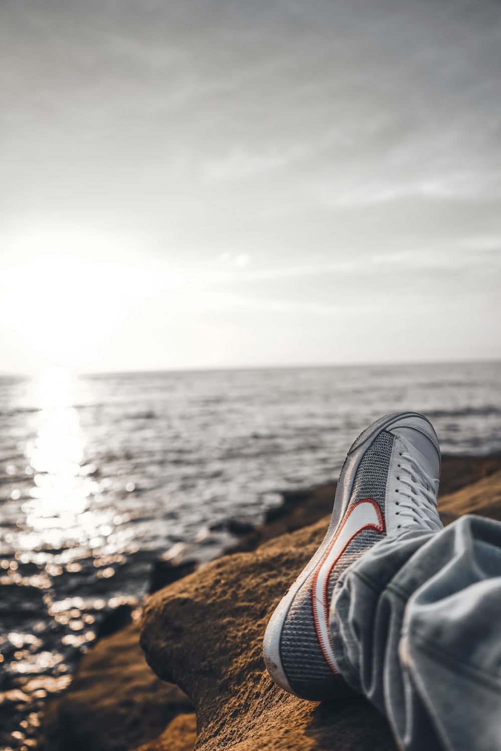 person in black and white sneakers sitting on brown rock near body of water during daytime