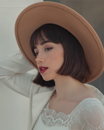 A woman with short dark hair is wearing a wide-brimmed tan hat and a lace-trimmed white top. Her makeup is subtle, with a focus on bold red lipstick and defined eyeliner. The lighting is soft, creating a gentle and elegant atmosphere.