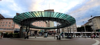 A public transportation station is depicted, featuring a modern circular glass and metal canopy in the center, surrounded by multiple tram tracks. The architecture of the surrounding buildings reflects an urban setting, with pedestrians moving around the area. Trams are visible on the right, with people waiting or walking nearby, suggesting a busy city atmosphere.