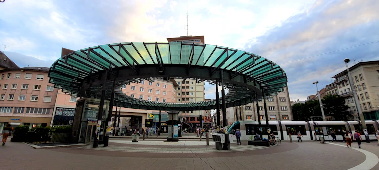 A public transportation station is depicted, featuring a modern circular glass and metal canopy in the center, surrounded by multiple tram tracks. The architecture of the surrounding buildings reflects an urban setting, with pedestrians moving around the area. Trams are visible on the right, with people waiting or walking nearby, suggesting a busy city atmosphere.