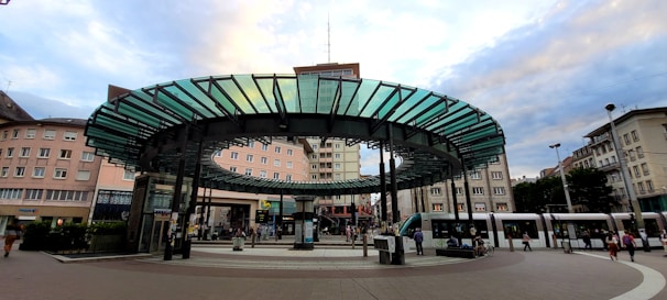 A public transportation station is depicted, featuring a modern circular glass and metal canopy in the center, surrounded by multiple tram tracks. The architecture of the surrounding buildings reflects an urban setting, with pedestrians moving around the area. Trams are visible on the right, with people waiting or walking nearby, suggesting a busy city atmosphere.