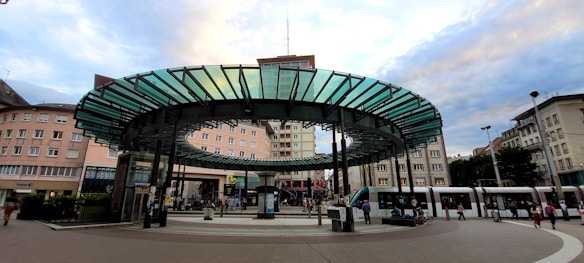 A public transportation station is depicted, featuring a modern circular glass and metal canopy in the center, surrounded by multiple tram tracks. The architecture of the surrounding buildings reflects an urban setting, with pedestrians moving around the area. Trams are visible on the right, with people waiting or walking nearby, suggesting a busy city atmosphere.