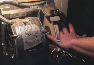 A close-up view of a hand dangerously close to a grinding wheel, with sparks flying off as the wheel spins. There is a diagram of a hand with fingers cut off taped to the grinding machine, possibly indicating the danger of using it. A hose with a metal cover can be seen attached to the machine.