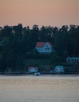 A serene landscape with a large house on a forested hill overlooking a body of water. Smaller houses are situated by the water's edge, and a boat can be seen gliding across the calm surface. The sky has a warm, dim glow, possibly indicating sunrise or sunset.