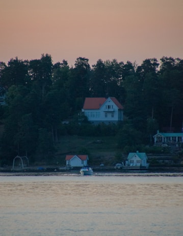 A serene landscape with a large house on a forested hill overlooking a body of water. Smaller houses are situated by the water's edge, and a boat can be seen gliding across the calm surface. The sky has a warm, dim glow, possibly indicating sunrise or sunset.