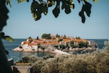 A picturesque island village sits surrounded by calm, blue sea. The island is covered with charming stone buildings topped with vibrant orange-red roofs, nestled among lush greenery. In the foreground, green leaves frame the view, adding to the serene and natural atmosphere.