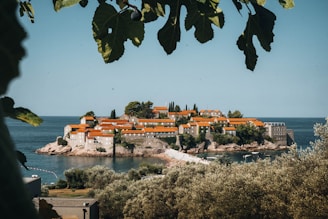 A picturesque island village sits surrounded by calm, blue sea. The island is covered with charming stone buildings topped with vibrant orange-red roofs, nestled among lush greenery. In the foreground, green leaves frame the view, adding to the serene and natural atmosphere.
