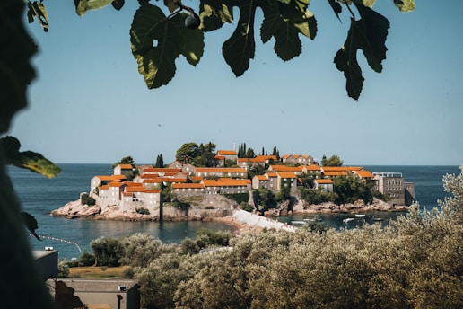 A picturesque island village sits surrounded by calm, blue sea. The island is covered with charming stone buildings topped with vibrant orange-red roofs, nestled among lush greenery. In the foreground, green leaves frame the view, adding to the serene and natural atmosphere.