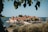 A picturesque island village sits surrounded by calm, blue sea. The island is covered with charming stone buildings topped with vibrant orange-red roofs, nestled among lush greenery. In the foreground, green leaves frame the view, adding to the serene and natural atmosphere.