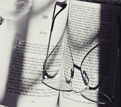 An open book displaying text on two pages with a pair of reading glasses resting on top. The shadows of the glasses are cast onto the pages, creating an artistic effect. The book appears to be a novel, and part of the page number and chapter title are visible.