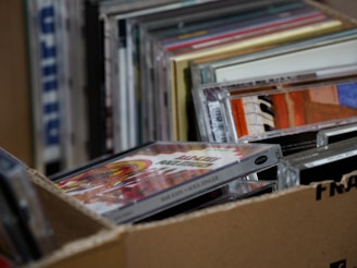 A collection of stacked CD cases inside a cardboard box, with visible album art and text on the covers. The CDs appear to be packed closely together, suggesting they are part of a larger collection.