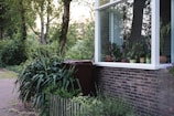 A serene garden scene featuring a brick house with a large window. Several potted plants are displayed on the window sill, and lush greenery surrounds the area, including tall trees and bushes. A brown recycling bin is placed near a small wooden fence bordering the garden.