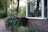 A serene garden scene featuring a brick house with a large window. Several potted plants are displayed on the window sill, and lush greenery surrounds the area, including tall trees and bushes. A brown recycling bin is placed near a small wooden fence bordering the garden.