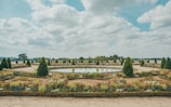 A beautifully landscaped garden featuring geometric layouts with neatly trimmed hedges, colorful flower beds, and symmetrical pathways. A circular water feature is centrally placed, mirroring the blue sky and scattered clouds. People are seen strolling around, suggesting a leisurely atmosphere.