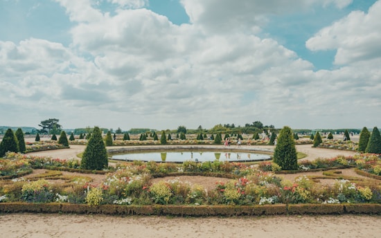 A beautifully landscaped garden featuring geometric layouts with neatly trimmed hedges, colorful flower beds, and symmetrical pathways. A circular water feature is centrally placed, mirroring the blue sky and scattered clouds. People are seen strolling around, suggesting a leisurely atmosphere.