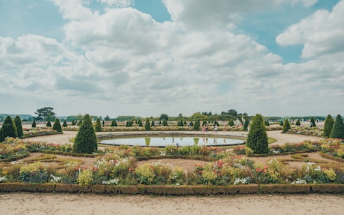 A beautifully landscaped garden featuring geometric layouts with neatly trimmed hedges, colorful flower beds, and symmetrical pathways. A circular water feature is centrally placed, mirroring the blue sky and scattered clouds. People are seen strolling around, suggesting a leisurely atmosphere.
