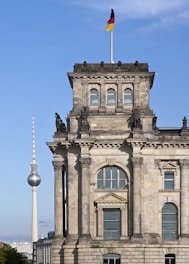 A historic stone building with ornate architecture features columns and arched windows. Above the building, a German flag waves on a pole, and in the background, the famous television tower rises against a clear blue sky.