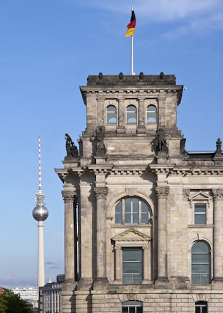 A historic stone building with ornate architecture features columns and arched windows. Above the building, a German flag waves on a pole, and in the background, the famous television tower rises against a clear blue sky.