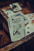 A playing card featuring a joker with a Las Vegas logo is prominently positioned among other scattered cards. The card has a vintage, worn look, resting against a rustic wooden surface. Various playing cards, partially visible, are scattered around with visible suits such as hearts and clubs.