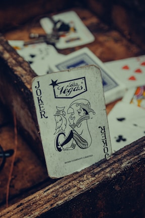 A playing card featuring a joker with a Las Vegas logo is prominently positioned among other scattered cards. The card has a vintage, worn look, resting against a rustic wooden surface. Various playing cards, partially visible, are scattered around with visible suits such as hearts and clubs.
