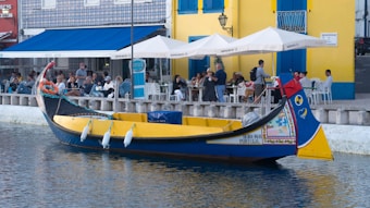 A colorful traditional boat is docked near the waterfront of a lively caf&eacute; area. People are seated at outdoor tables beneath white umbrellas, enjoying their time and engaging in conversations. The buildings in the background are painted in vibrant colors like yellow and blue, further adding to the cheerful ambiance of the scene.