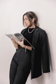 A person is leaning against a pale wall while reading a magazine. They are dressed in a stylish black suit and wearing jewelry, including earrings and bracelets. Their hair is tied back neatly, and the expression appears thoughtful and focused.