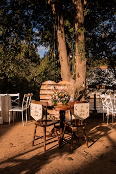 Outdoor setting with two wooden chairs facing a round table topped with a colorful floral arrangement, set against a large tree. White chairs and a table are in the background, with warm sunlight casting shadows on the sandy ground. Hanging light bulbs are visible among the tree branches, adding charm to the rustic atmosphere.