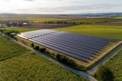 An expansive solar panel farm is situated in a rural landscape, surrounded by vast green fields and distant hills. The neatly arranged rows of solar panels reflect the sunlight, contrasting with the lush, grassy terrain. A few scattered trees and agricultural buildings can be seen in the background.
