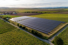 An expansive solar panel farm is situated in a rural landscape, surrounded by vast green fields and distant hills. The neatly arranged rows of solar panels reflect the sunlight, contrasting with the lush, grassy terrain. A few scattered trees and agricultural buildings can be seen in the background.