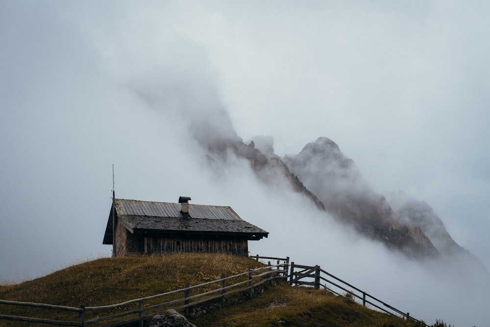Braunes Holzhaus auf grünem Grasfeld in der Nähe von Bergen, die tagsüber mit Nebel bedeckt sind