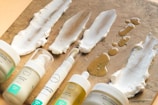 Various skincare products in pump bottles and tubs are arranged on a textured brown surface, with some of the creams and oils spread in swatches on the surface.