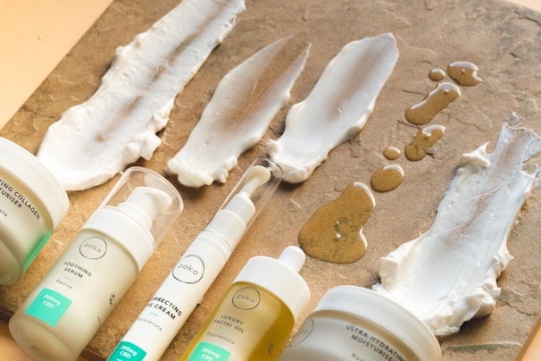 Various skincare products in pump bottles and tubs are arranged on a textured brown surface, with some of the creams and oils spread in swatches on the surface.